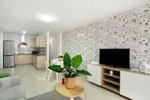 Una cocina moderna con estufa integrada, un comedor con mesa y sillas blancas, una sala de estar con televisor y mueble de entretenimiento, y una planta grande en maceta.