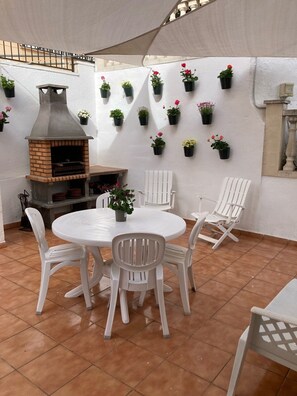 An outdoor patio with a brick oven, white plastic chairs, a round table, and potted plants on the wall. - Holiday Home "Lola's Beach House" with Sea View, Private Pool & Wi-Fi (Calafell)