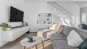 Ein modernes Wohnzimmer mit einem grauen Sofa, einem Flachbildfernseher, einem weißen Konsolentisch mit Pflanzen und einer Treppe.