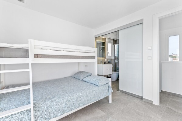 A bunk bed with a patterned bedspread, a white wardrobe, a bathroom with a glass shower, and a door leading to another room.