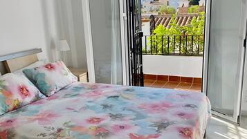 A bedroom with a bed, a nightstand, a lamp, and a view of a balcony with a railing.