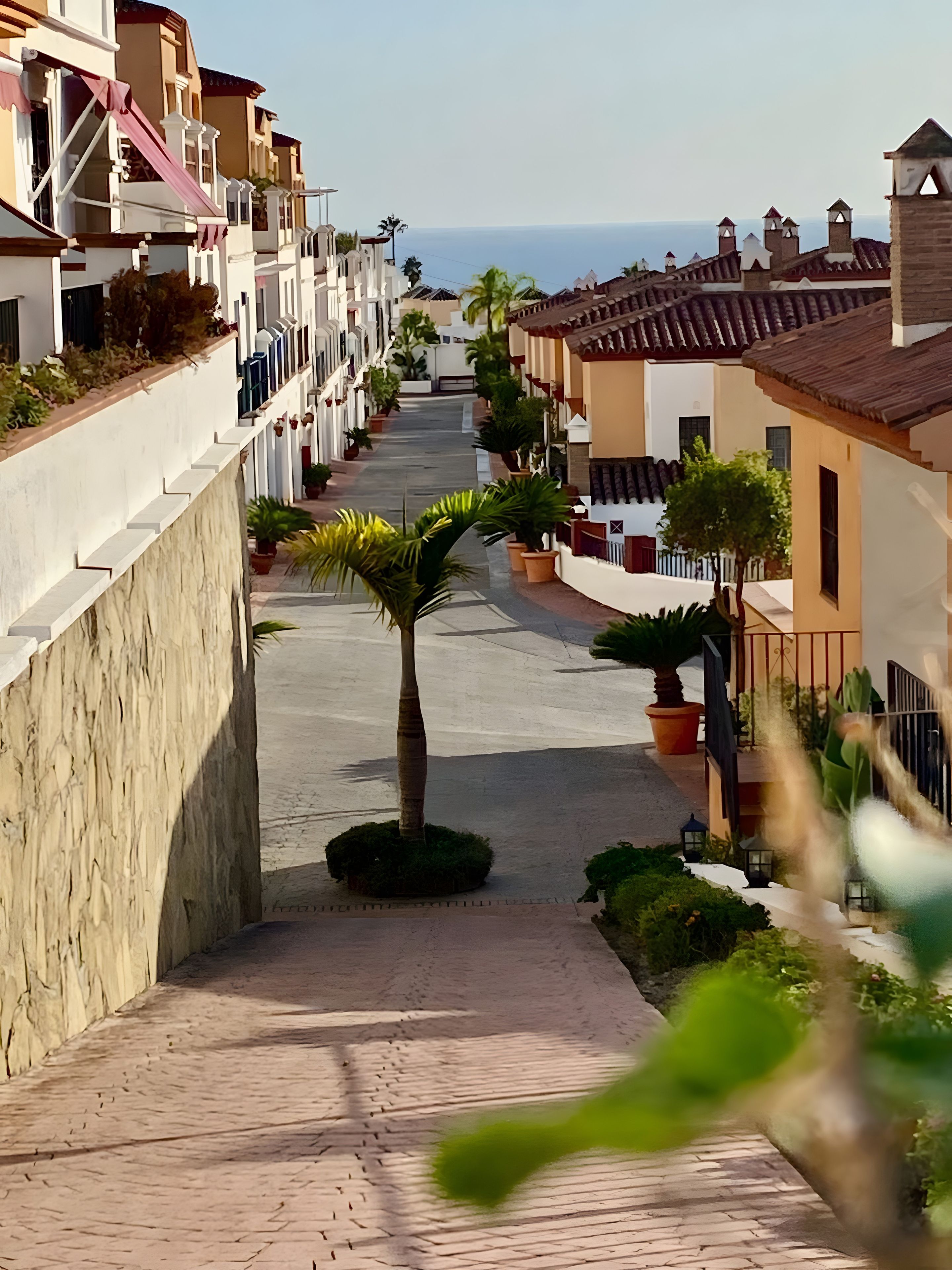 A residential street with houses on both sides, a central pathway, and a view of the sea in the distance.