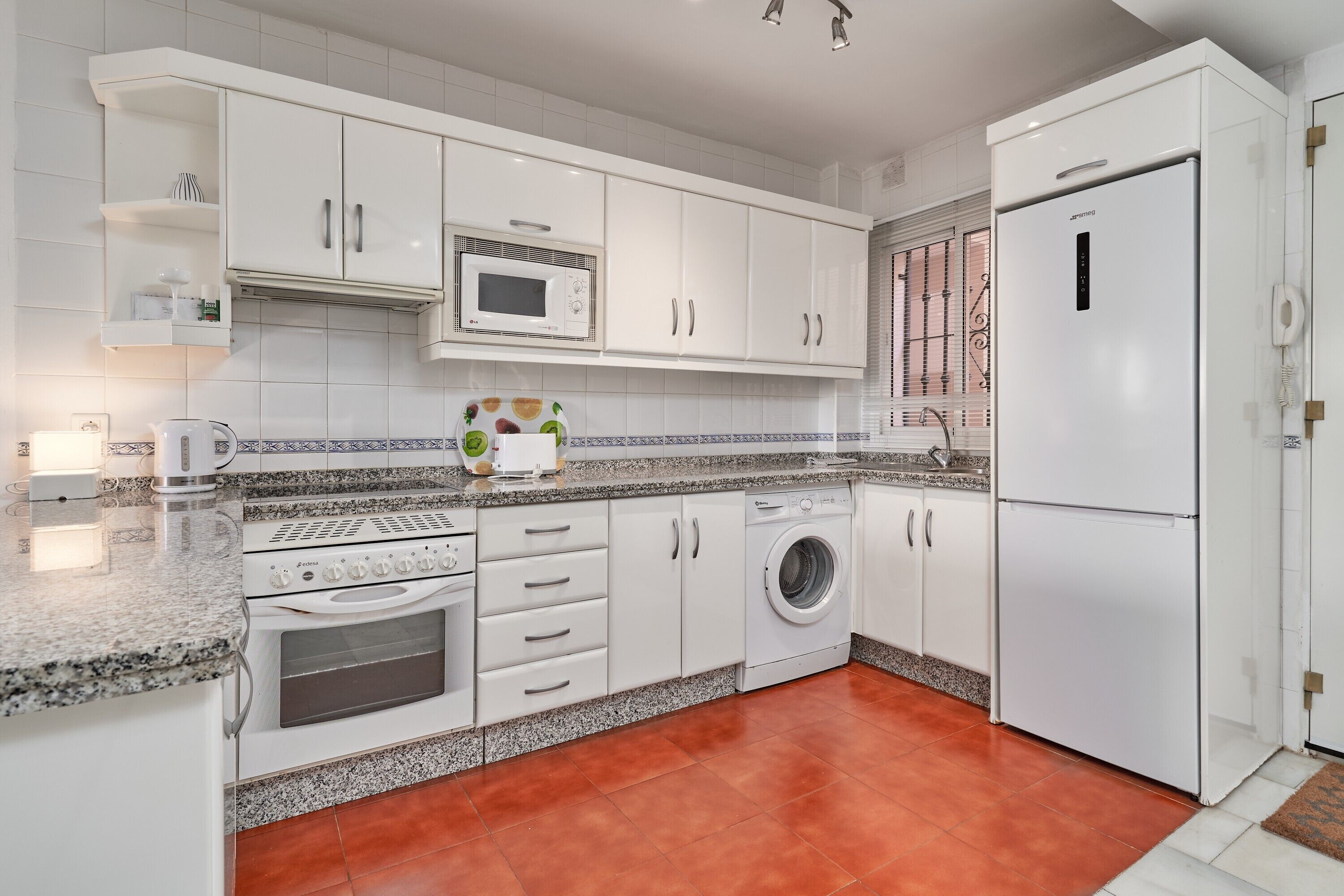 Uma cozinha moderna com armários brancos, micro-ondas, máquina de lavar e frigorífico.