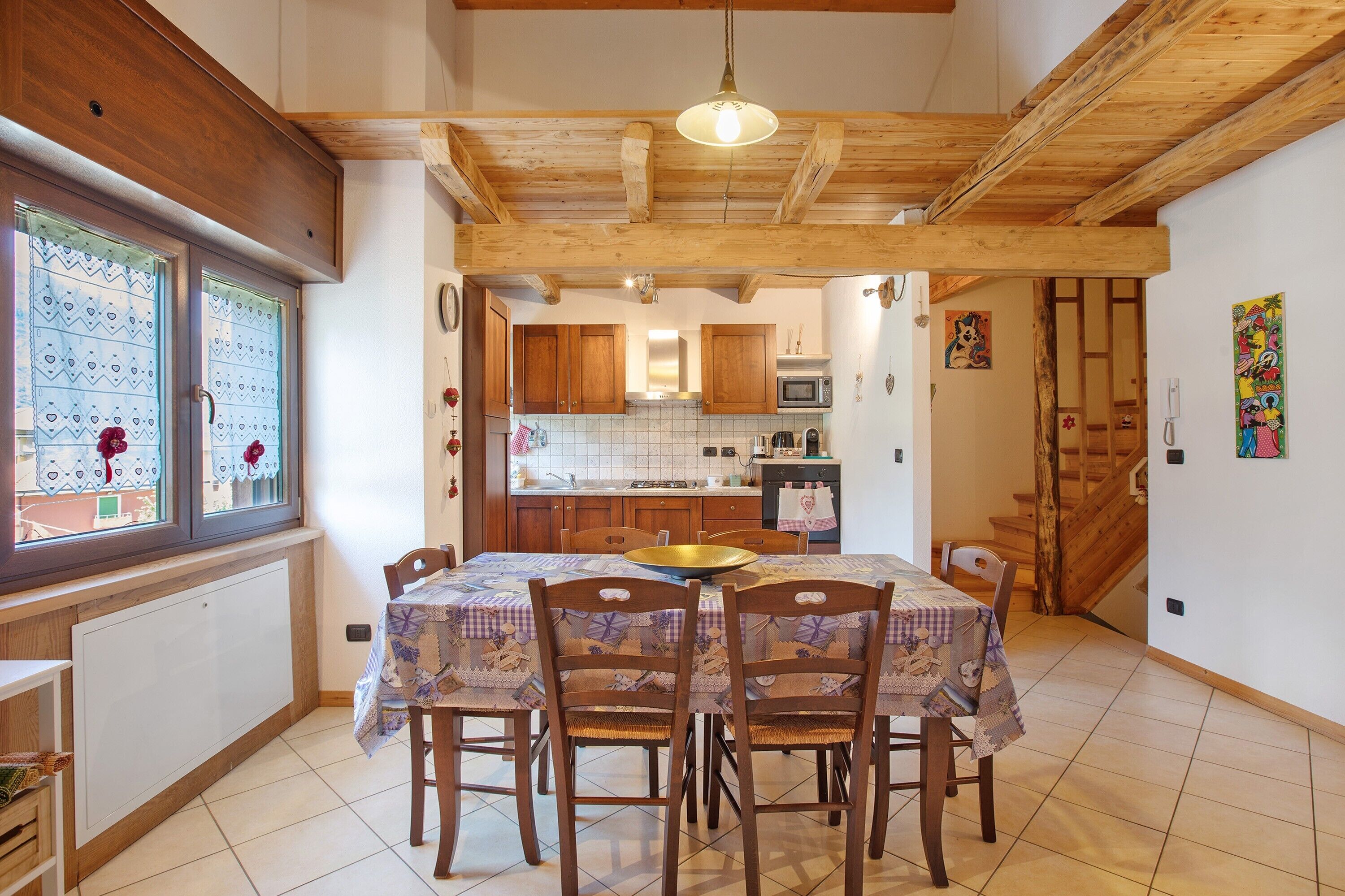 A dining area with a table set for four, wooden chairs, a hanging light fixture, and a view through a window with patterned curtains.