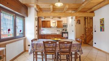 A dining area with a table set for four, wooden chairs, a hanging light fixture, and a view through a window with patterned curtains.