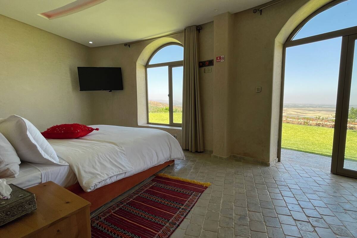 A bedroom with a bed, a TV mounted on the wall, and large windows offering a view of the countryside.