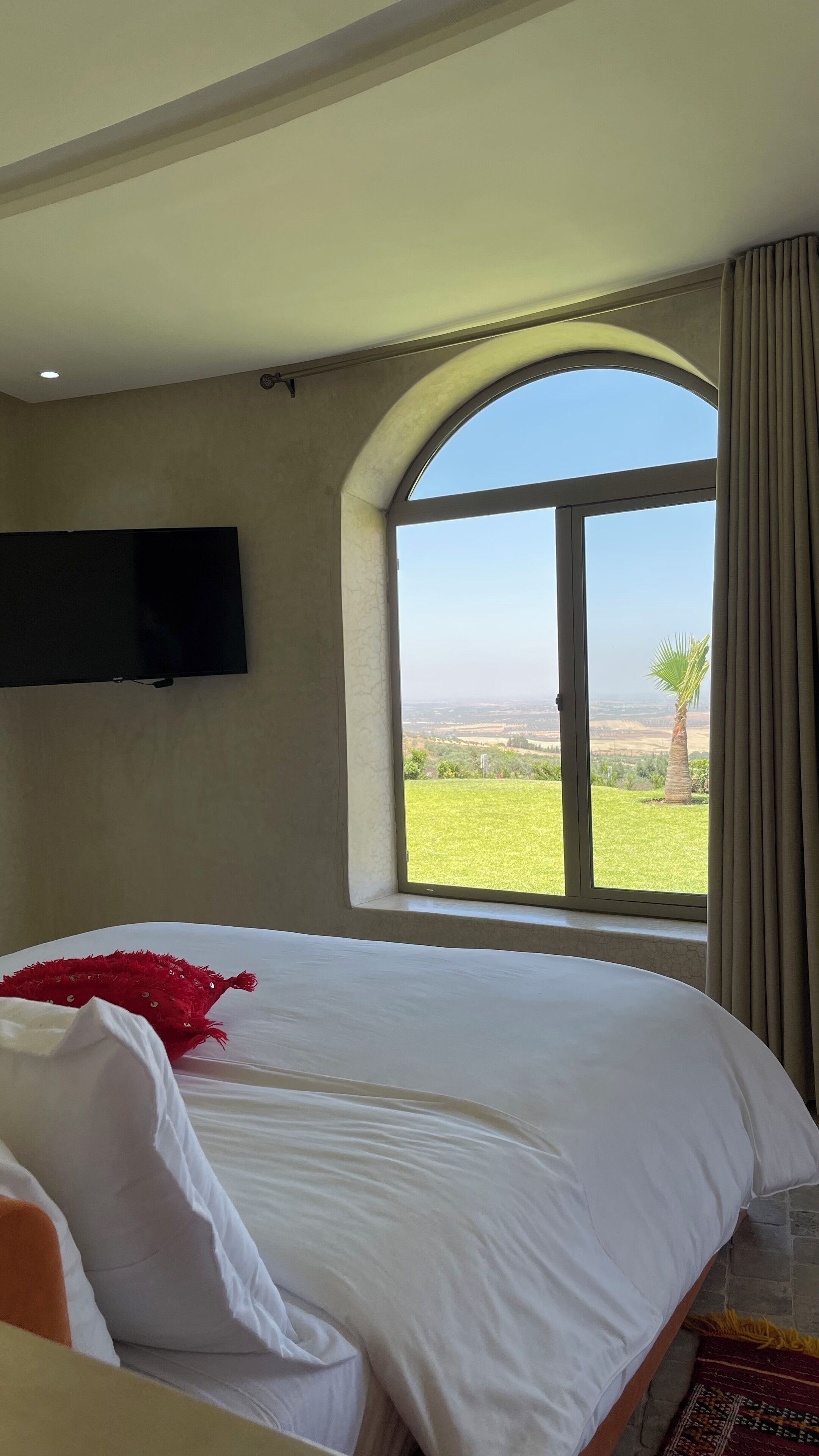 A hotel room with a large bed, a flat-screen TV mounted on the wall, and a large arched window offering a view of the countryside.