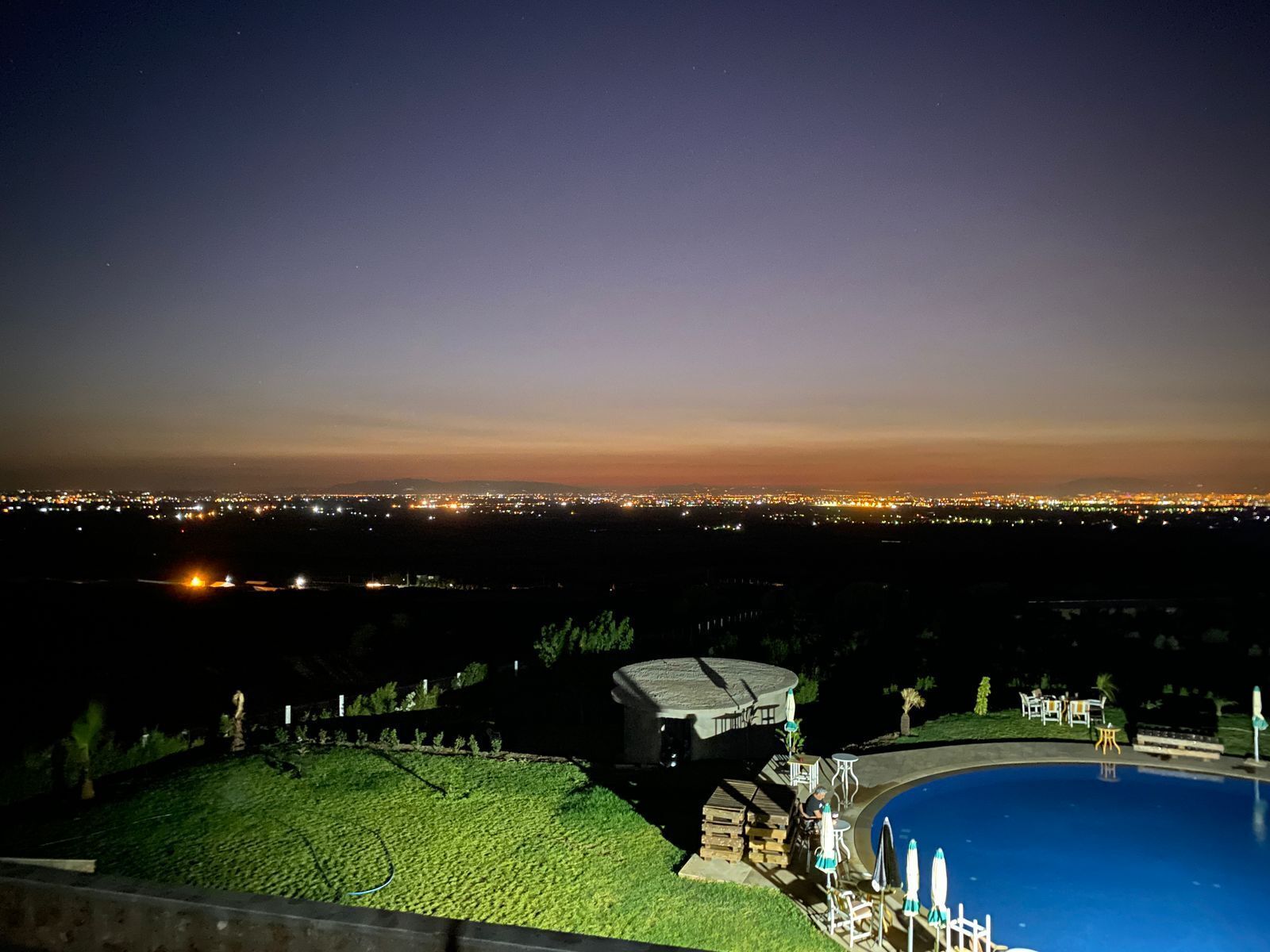 A nighttime view of a residential area with a swimming pool, a covered patio, and a distant cityscape.