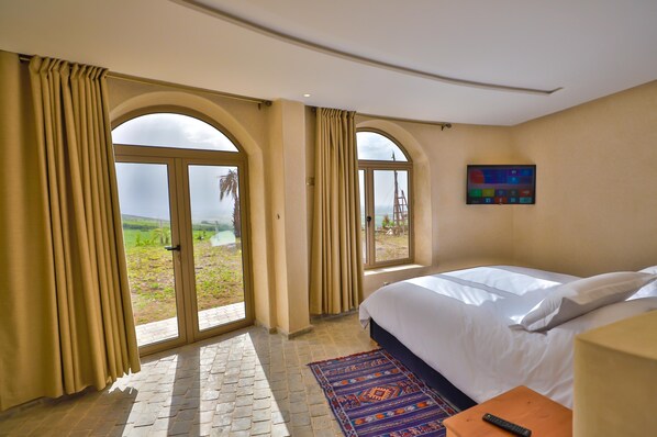 A hotel room with a large bed, a TV on the wall, and a view of the outdoors through arched windows. - Bhalil Farm House (Bhalil)
