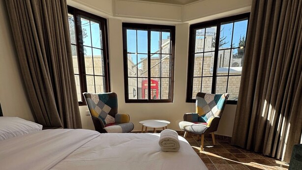 A hotel room with a large bed, two armchairs, a small table, and a view of a building through large windows. - 1 Antika Jabalal-lovebdeh (Amman)