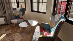 A room with two patterned armchairs, a small white table, and large windows with a view of a brick wall and a red telephone booth. - 1 Antika Jabalal-lovebdeh (Amman)