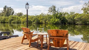 A wooden deck with two chairs and a small table, overlooking a calm lake with dense greenery and a lamp post.