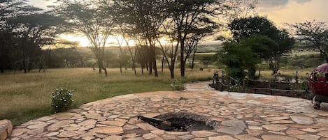 A stone patio with a fire pit, surrounded by a grassy field and trees, with a sunset in the background.