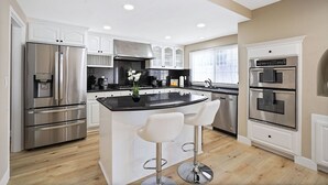 A modern kitchen with stainless steel appliances, white cabinetry, and a central island with bar stools. - Spacious Clean Home w/ Quality Furniture close to Disneyland & Huntington Beach! (Garden Grove)