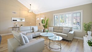 A spacious living room with a high ceiling, a central light fixture, two sofas, a coffee table, and large windows with blinds. - Spacious Clean Home w/ Quality Furniture close to Disneyland & Huntington Beach! (Garden Grove)