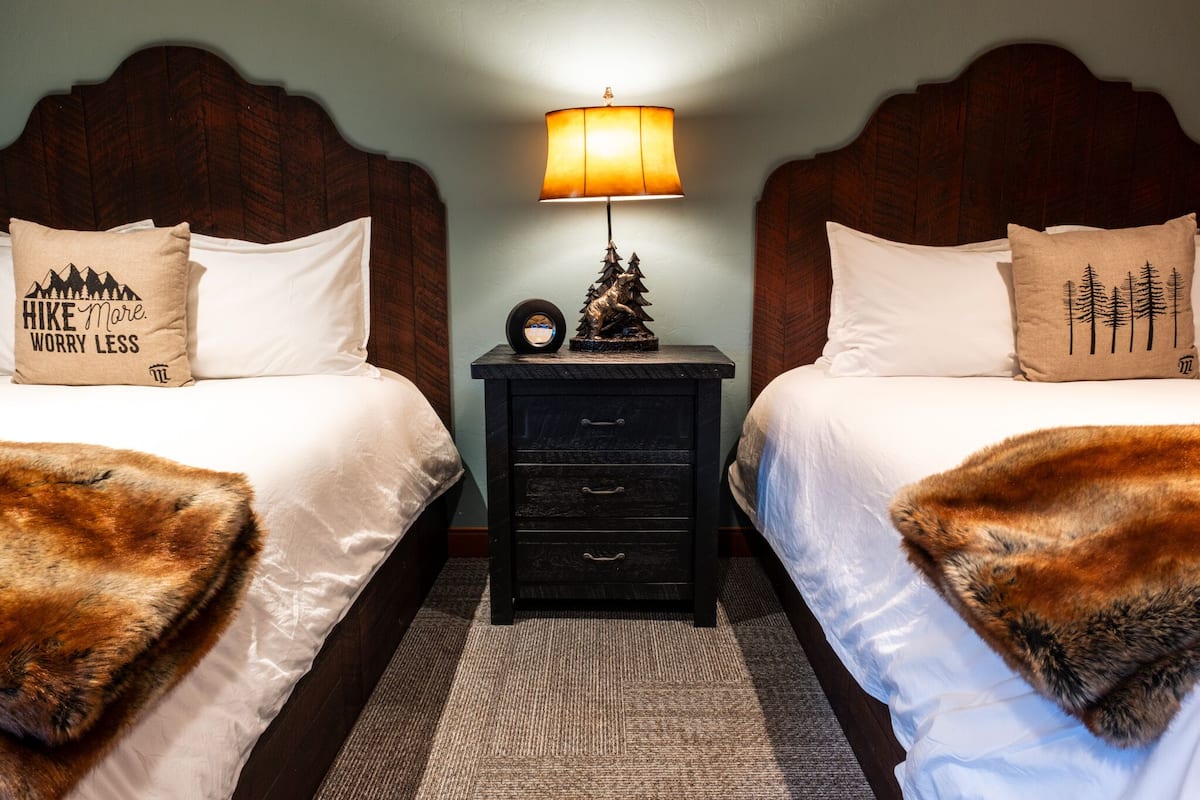 Een hotelkamer met twee bedden, elk met een houten hoofdbord, een nachtkastje met een lamp en decoratieve kussens.