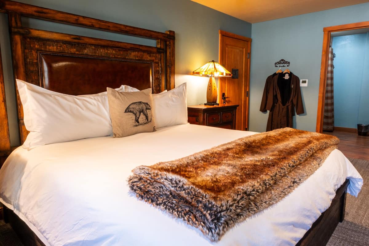 Een netjes opgemaakt bed met een witte deken en een bont vest, een houten hoofdbord, een lamp en een kapstok met een jas.