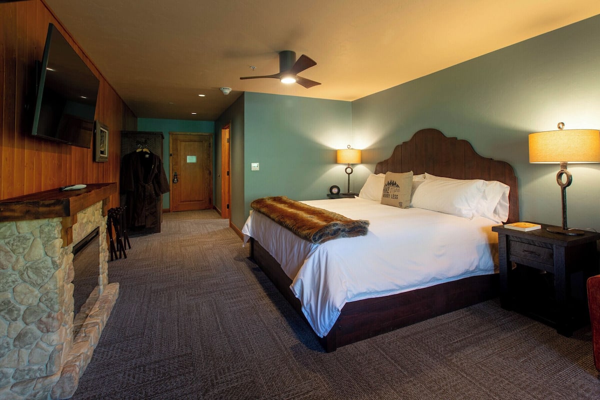 Een hotelkamer met een stenen openhaard, een bed met een witte deken, een houten hoofdbord, een plafondventilator en twee bedlampen.