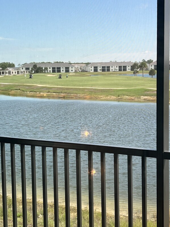 A view of a golf course with a body of water in the foreground, a wooden deck, and a row of buildings in the background.