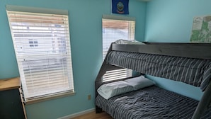 Un dormitorio con literas, suelo de madera, un pequeño banco y dos ventanas con persianas.