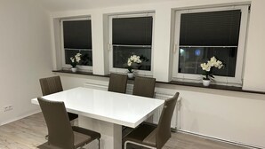 A modern dining area with a white table and brown chairs, three windows with blinds, and potted white flowers on the windowsills. - Wohnen wie zu Hause (Löhne)