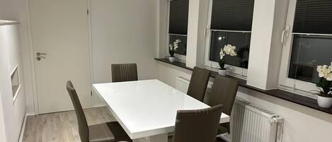A dining area with a white table and four brown chairs. There are three windows with blinds, each with a potted plant on the windowsill.