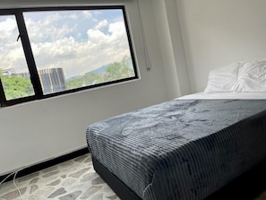 Ein Schlafzimmer mit Bett, Fenster und Blick auf Gebäude und Grünflächen.