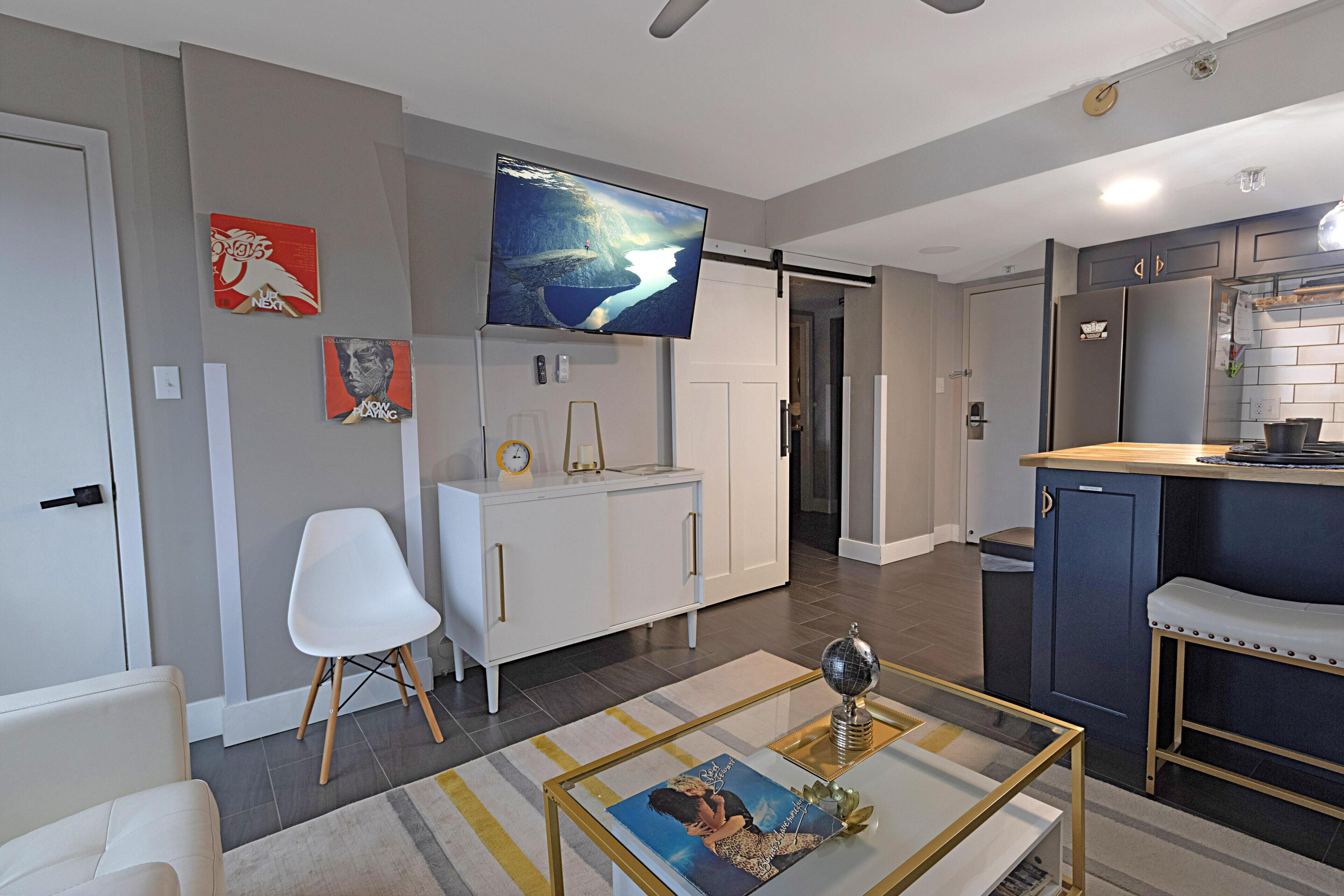 A modern living room with a flat-screen TV, white furniture, and a kitchen area with a blue countertop.