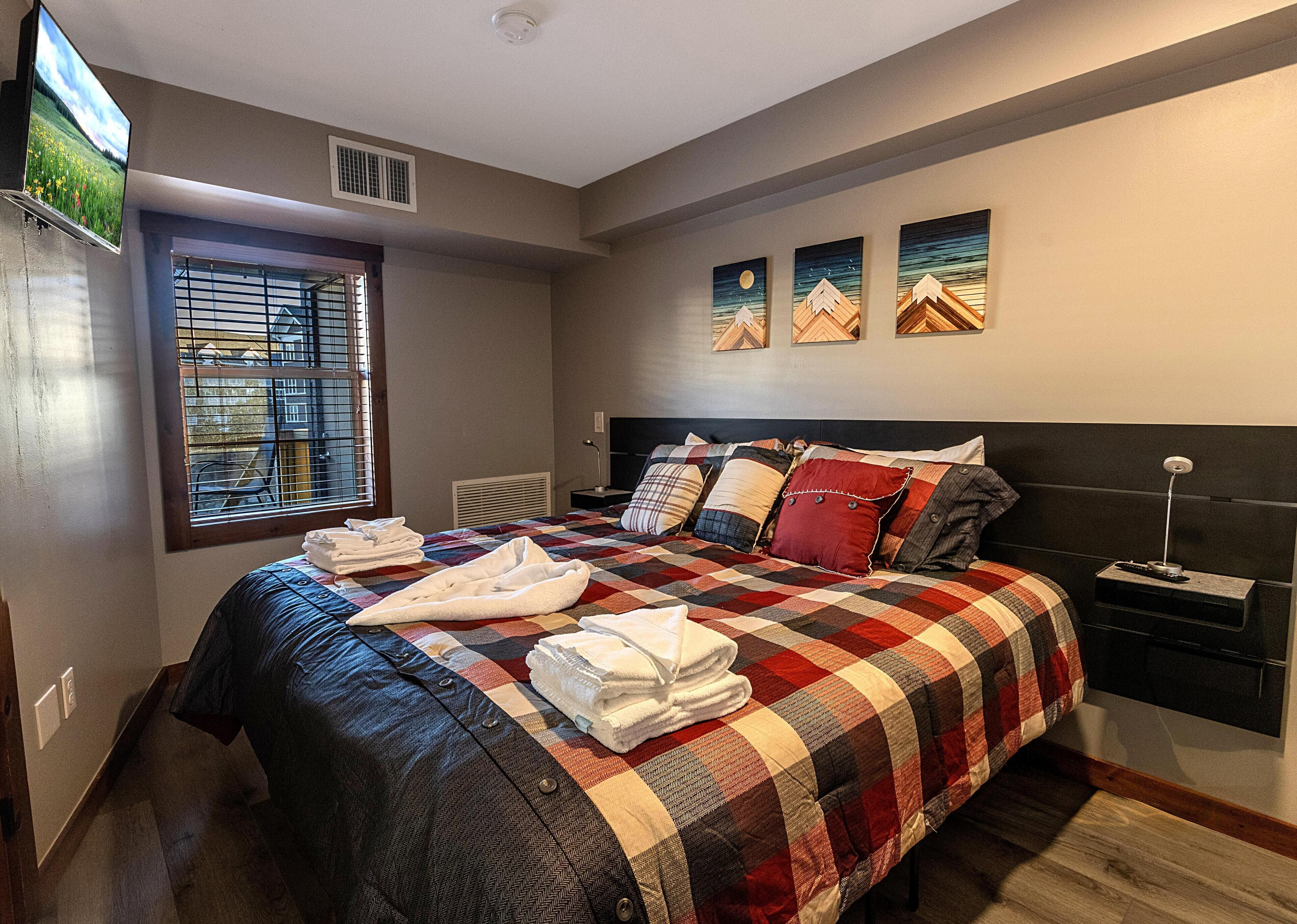 Una habitación de hotel con una cama grande, un televisor montado en la pared, una ventana con persianas y obras de arte en la pared.