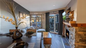 A modern living room with a stone fireplace, a sofa, a coffee table, and a dining table with a vase and bowls. - RL 419 - High-Elevation Renovated Summer Escape in Prime Village Spot (Snowshoe)