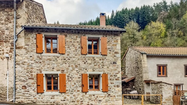 A stone building with wooden shutters, a chimney, and a view of a forested hillside.