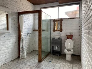 A small, rustic bathroom with a white brick wall, a glass shower enclosure, a wooden door, a white toilet, and a small sink with a mirror above it. - 5 de Mayo Boutique Loft (Tijuana)