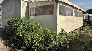 A small, single-story mobile home with a covered porch, surrounded by dense green foliage.