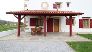 A covered patio with a stone floor, a wooden table and chairs, and a red door. - APEZAINEA - SARE - Gîte (SARE)
