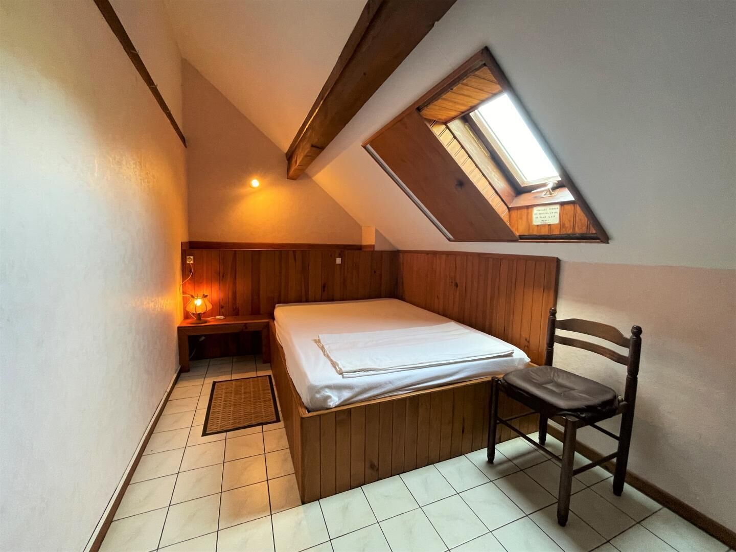 Une petite pièce agréable, avec un plafond en pente, un lit simple, une chaise, une table de chevet et un puits de lumière.