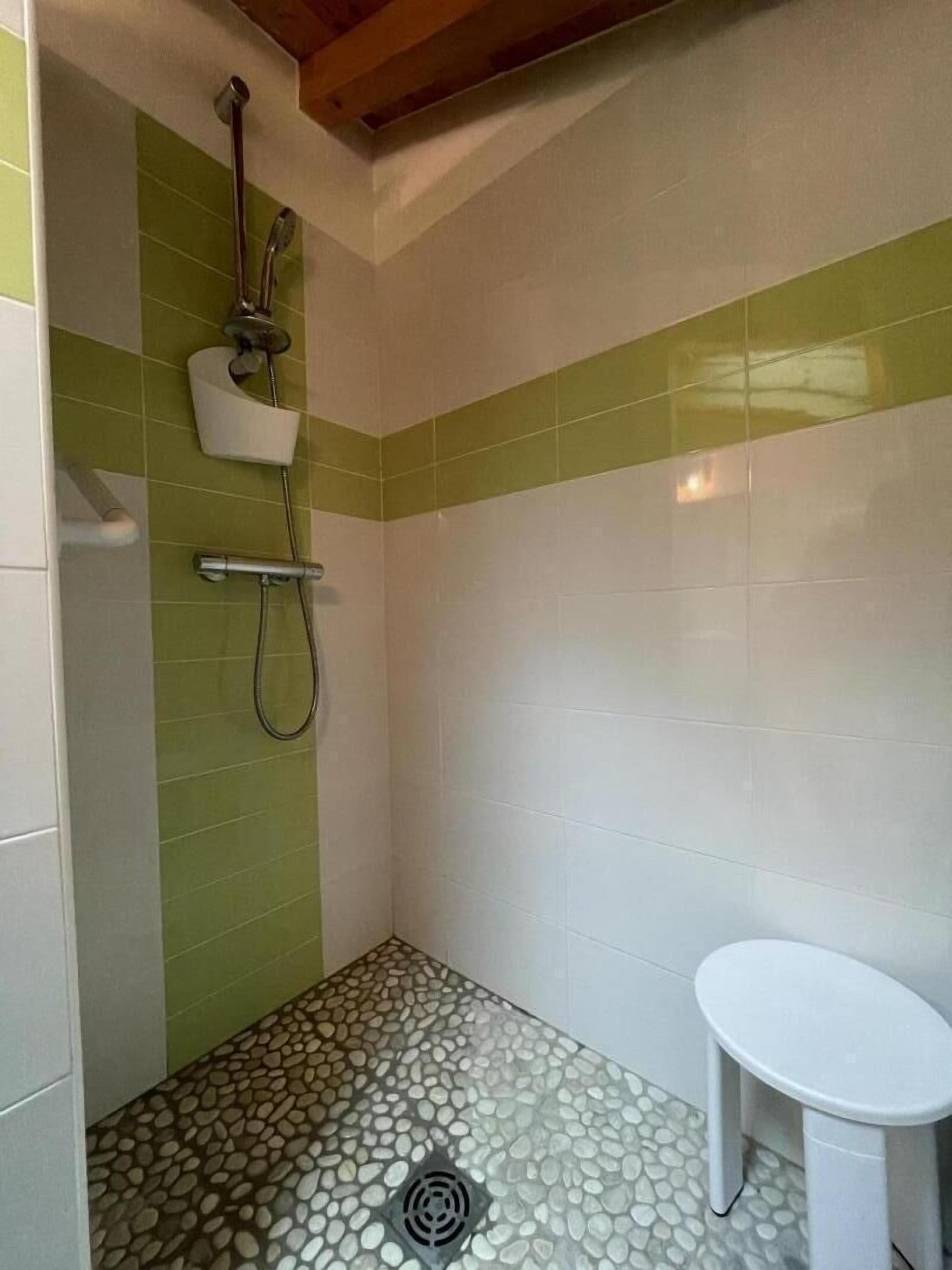 A small bathroom with green and white tiled walls, a shower with a handheld showerhead, a toilet, and a pebble-patterned floor.