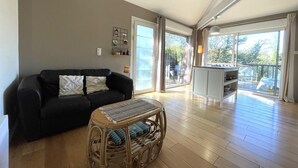 Ein großzügiger Wohnbereich mit einer schwarzen Couch, einem Korbtisch und Blick auf einen Balkon mit Holzboden.