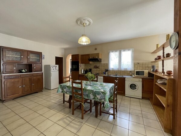 A kitchen with wooden cabinets, a dining table with chairs, a refrigerator, a washing machine, and a window with curtains.