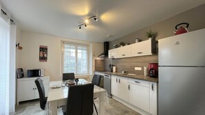 A modern kitchen with white cabinets, a stainless steel refrigerator, and a dining area with a table and chairs. - Uharte Beherea - Souraide (SOURAIDE)