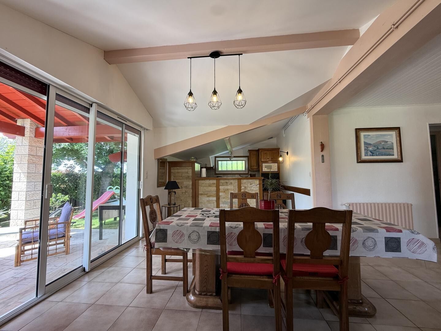 Uma área de refeições com uma mesa posta para quatro pessoas, cadeiras de madeira, uma porta de vidro deslizante que dá para uma área externa e uma cozinha visível através de uma abertura.