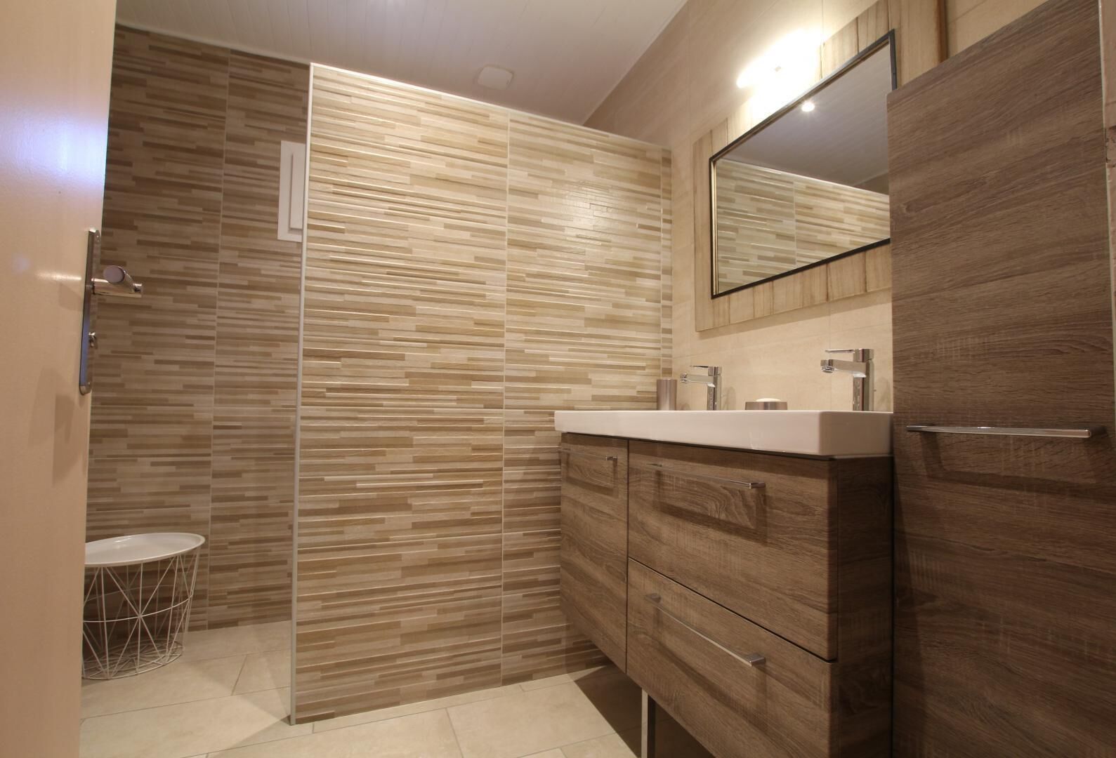A modern bathroom with a wooden vanity, a mirror, and a shower area with a tiled wall.