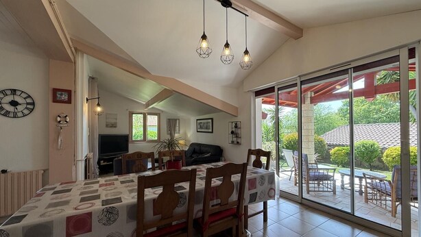 A dining area with a table set for four, a television in the background, and a view of the outdoors through a sliding glass door. - Xirimola BI - Saint-pee-sur-nivelle (SAINT-PEE-SUR-NIVELLE)
