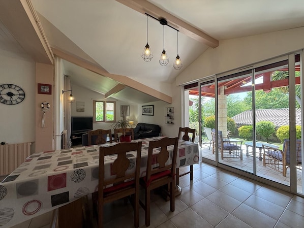 A dining area with a table set for four, a television in the background, and a view of the outdoors through a sliding glass door.