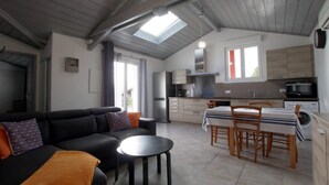 Een moderne woonkamer met een bank, een ronde tafel en een eethoek met stoelen. In de achtergrond is een keuken zichtbaar met een fornuis, magnetron en vaatwasser.