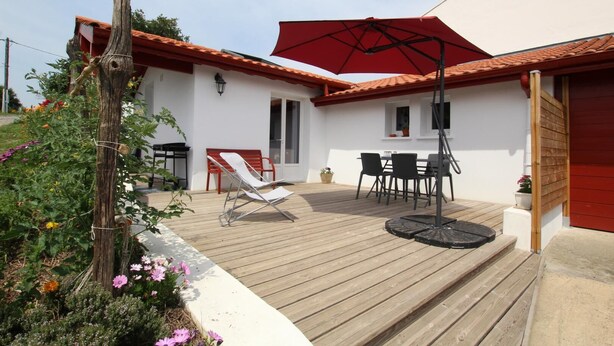 Een houten terras met een rode parasol, een witte stoel en een tafel met stoelen.