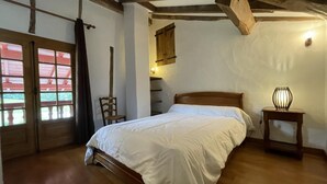 A bedroom with a wooden bed, a nightstand with a lamp, a chair, and a window with blinds. - Zamatelua 2 - Sare - Gîte (SARE)