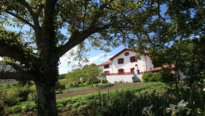 A white building with red accents, surrounded by lush greenery and a tree in the foreground. - Zamatelua 2 - Sare - Gîte (SARE)