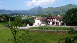 A traditional chalet-style building with a sloped roof, surrounded by lush green fields and mountains in the background. - Zamatelua 2 - Sare - Gîte (SARE)