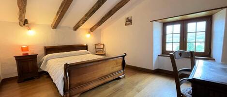A bedroom with a wooden bed, a nightstand, a desk with a chair, and a window with a view.