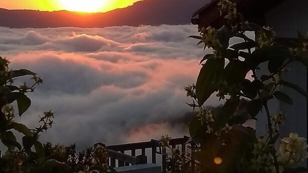 Un lever de soleil sur un paysage montagneux, avec des nuages en contrebas, et un bâtiment doté d'un balcon au premier plan.